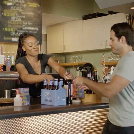 woman in black shirt and braids behind a counter hands alcoholic beverages to man in grey shirt with brown hair